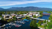 Aerial view of downtown Sandpoint, Idaho — the Sand Creek waterfront and marina, the Cedar Street Bridge, and mountains beyond